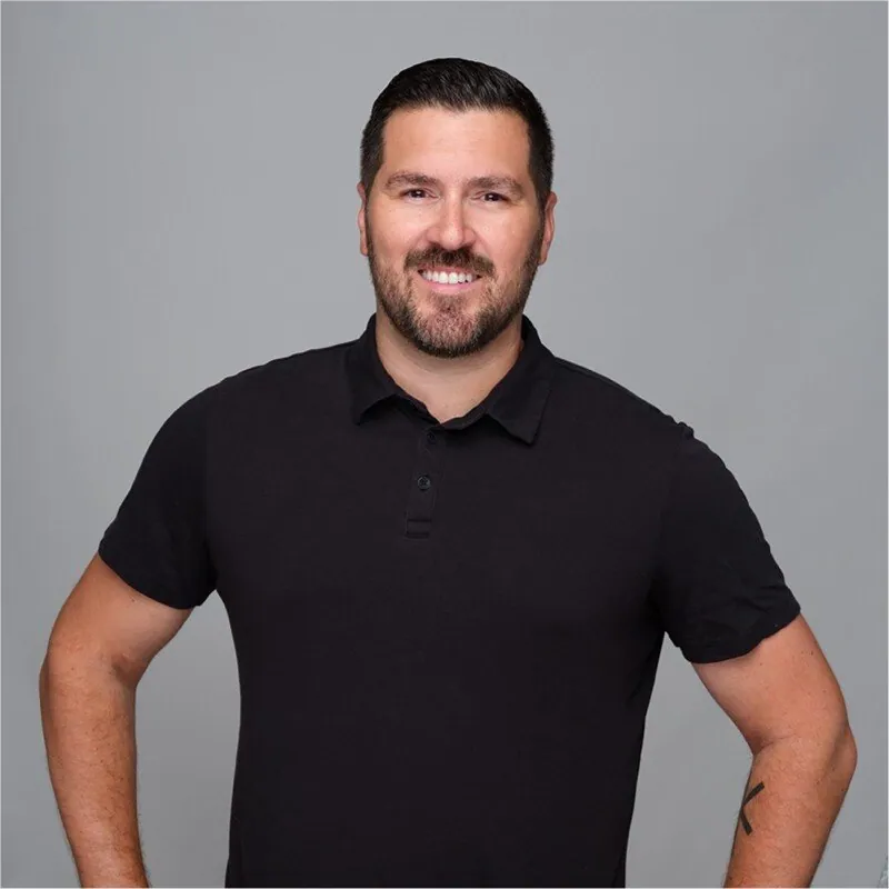 A man with a beard and short dark hair smiling, wearing a black polo shirt, standing against a gray background.