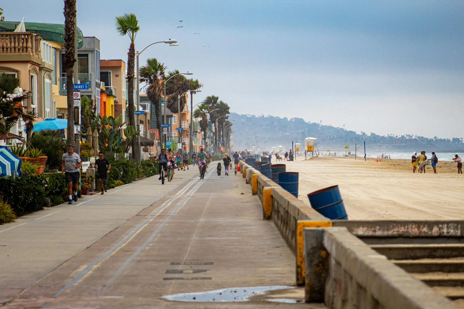 A lively beachfront promenade with people walking and biking, lined with colorful houses, palm trees, and street lamps, leading to the sandy beach and ocean in the distance on a cloudy day.