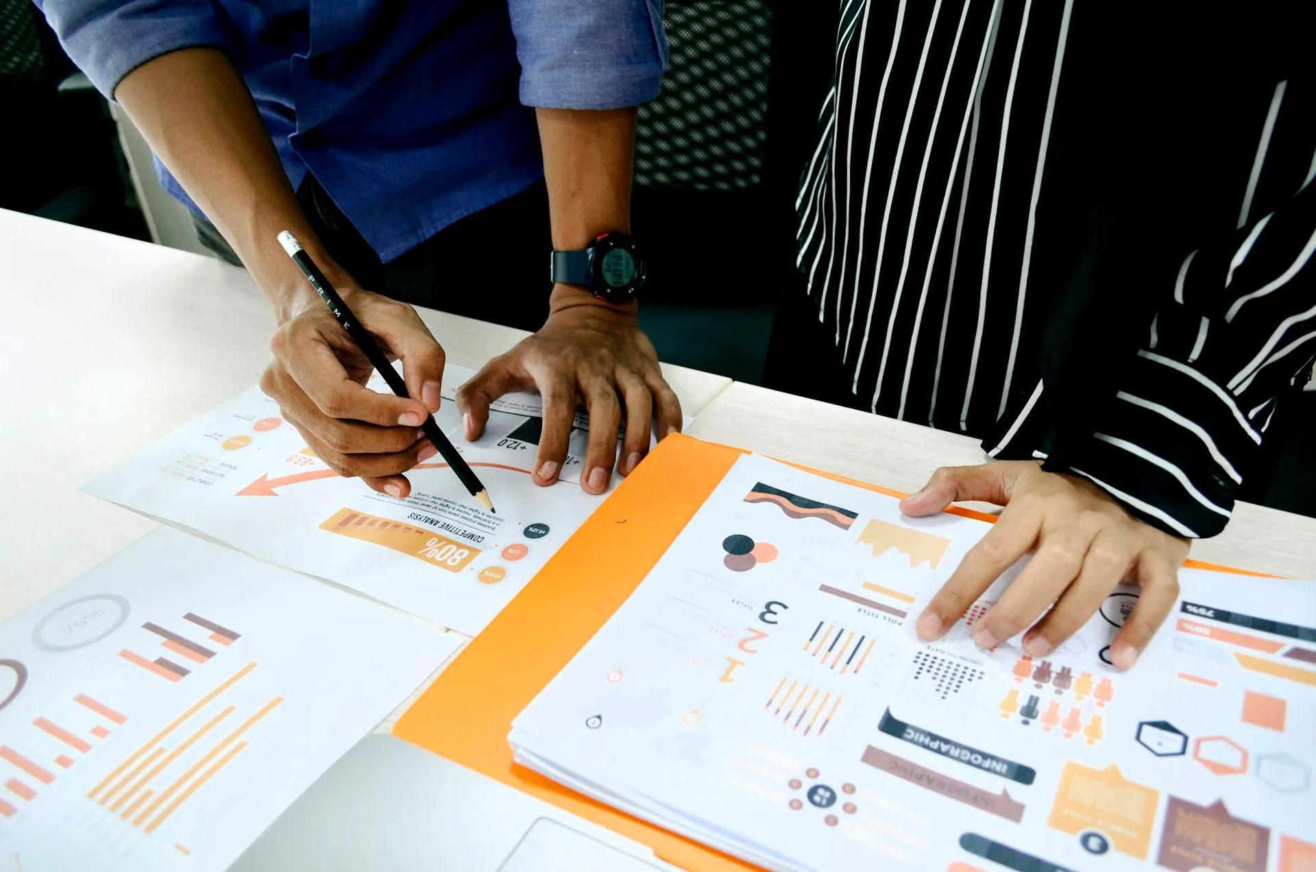 Two people discussing business reports and charts on a desk, with one pointing at a document with a pen.