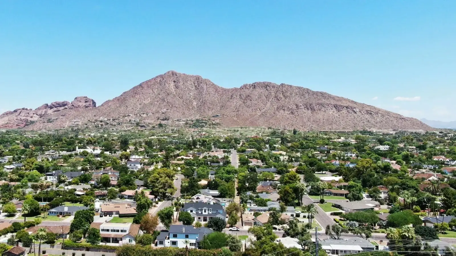 Aerial view of a suburban neighborhood with houses and trees in front of a large rocky mountain under a clear blue sky.