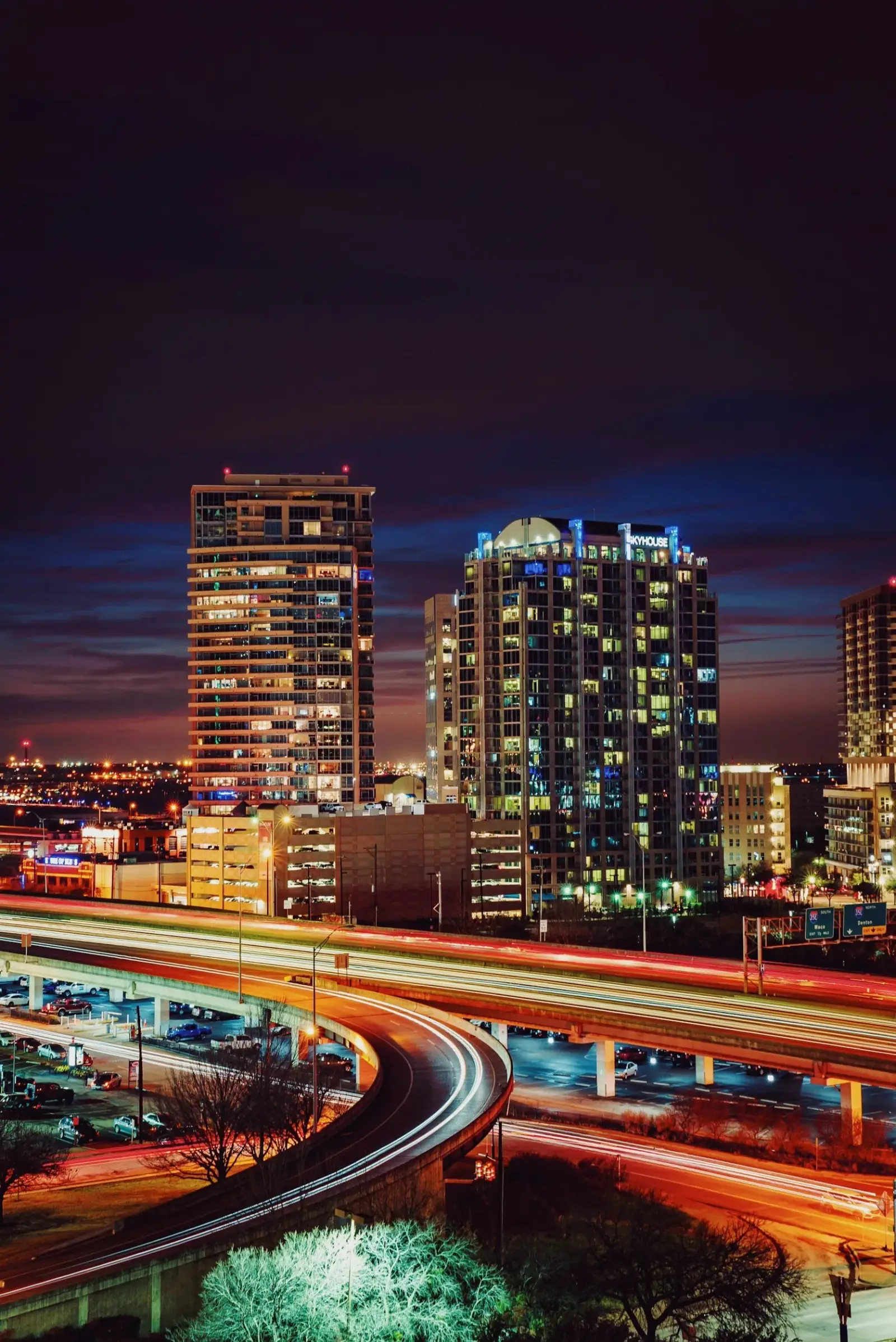 Nighttime cityscape with illuminated high-rise buildings, a highway with light trails from moving vehicles, and a dark sky with some clouds.