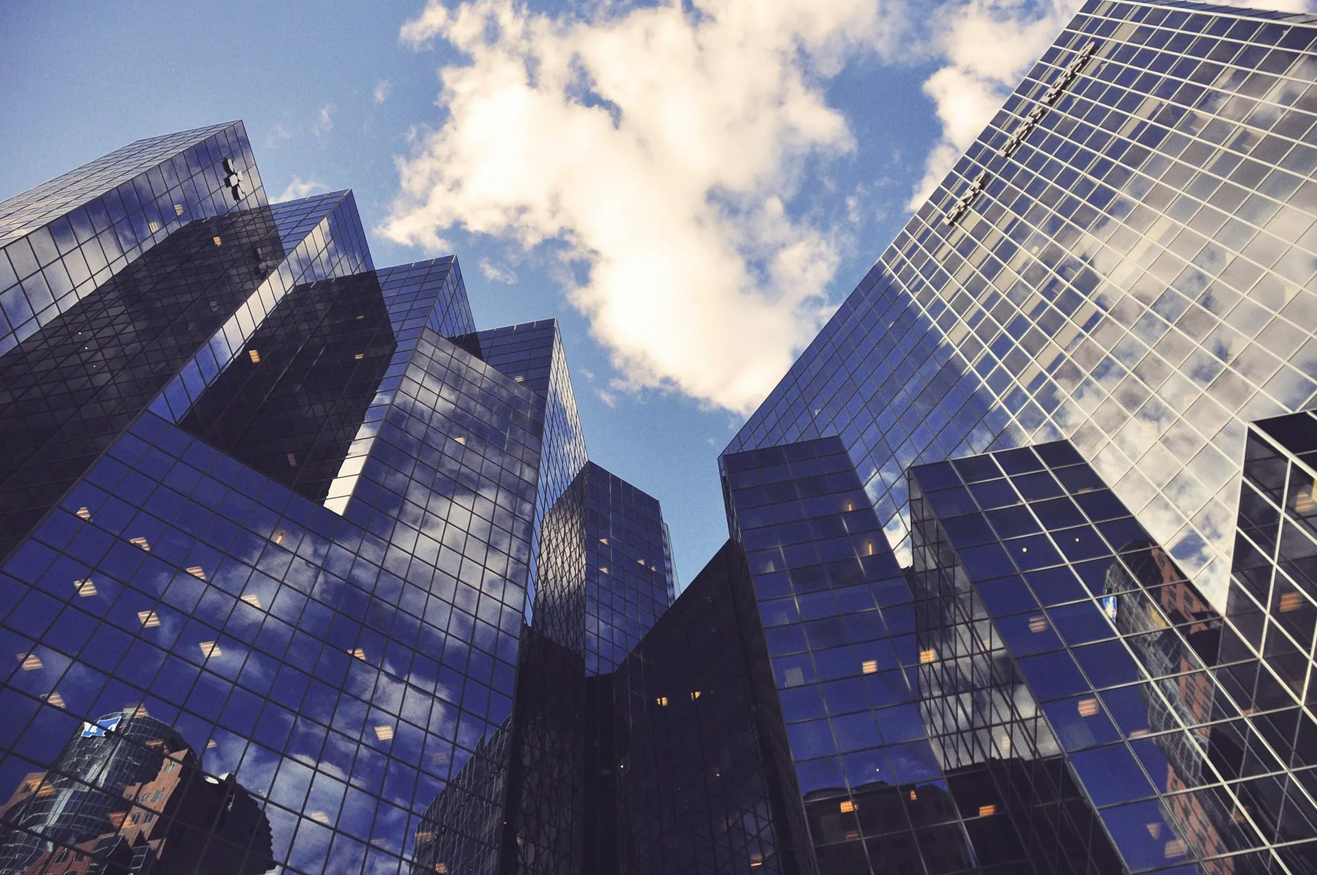 Reflections of clouds in the glass windows of tall modern skyscrapers against a daytime blue sky.