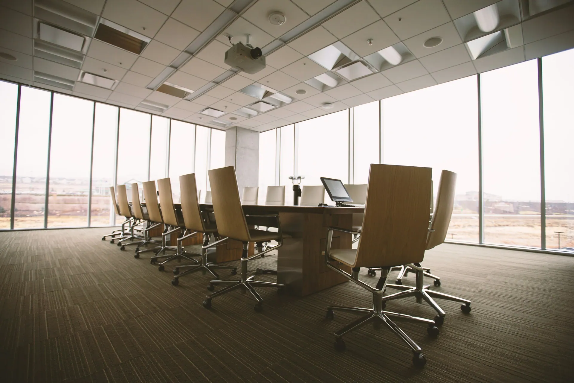 Empty modern conference room with large windows, a wooden table, beige chairs on casters, a projector, a laptop, and a view of the city and landscape outside.