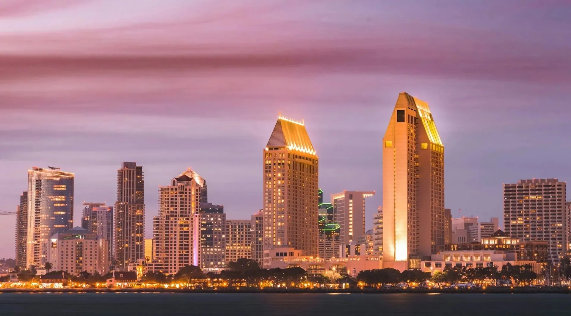 Nighttime skyline of downtown San Diego with illuminated high-rise buildings and purple clouded sky.