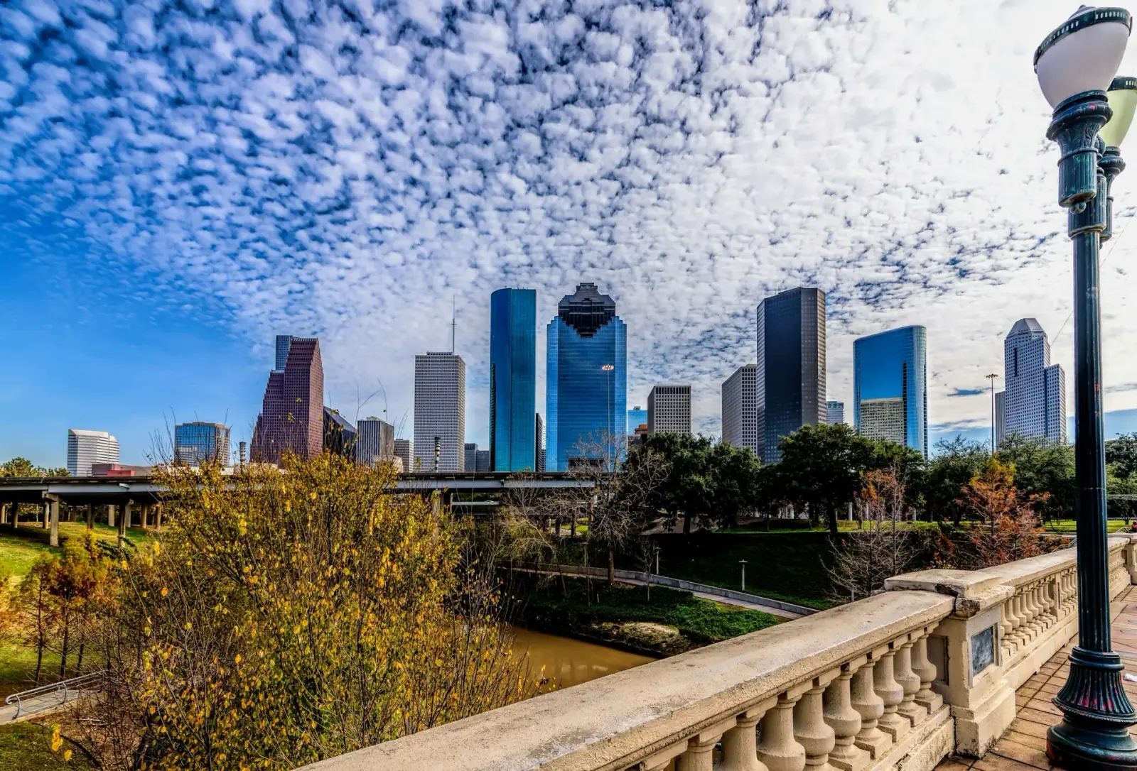 Downtown Houston skyline with skyscrapers and vibrant clouds in the sky, seen from a park with a stone railing and streetlamp in the foreground.