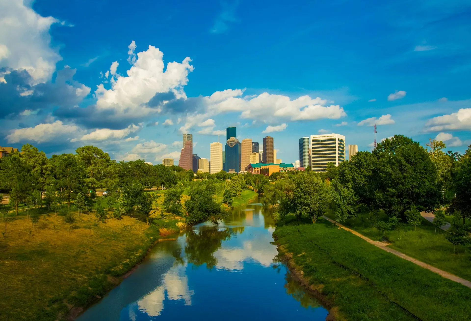 A city skyline with tall buildings in the distance and a river running through a green park in the foreground under a partly cloudy sky.