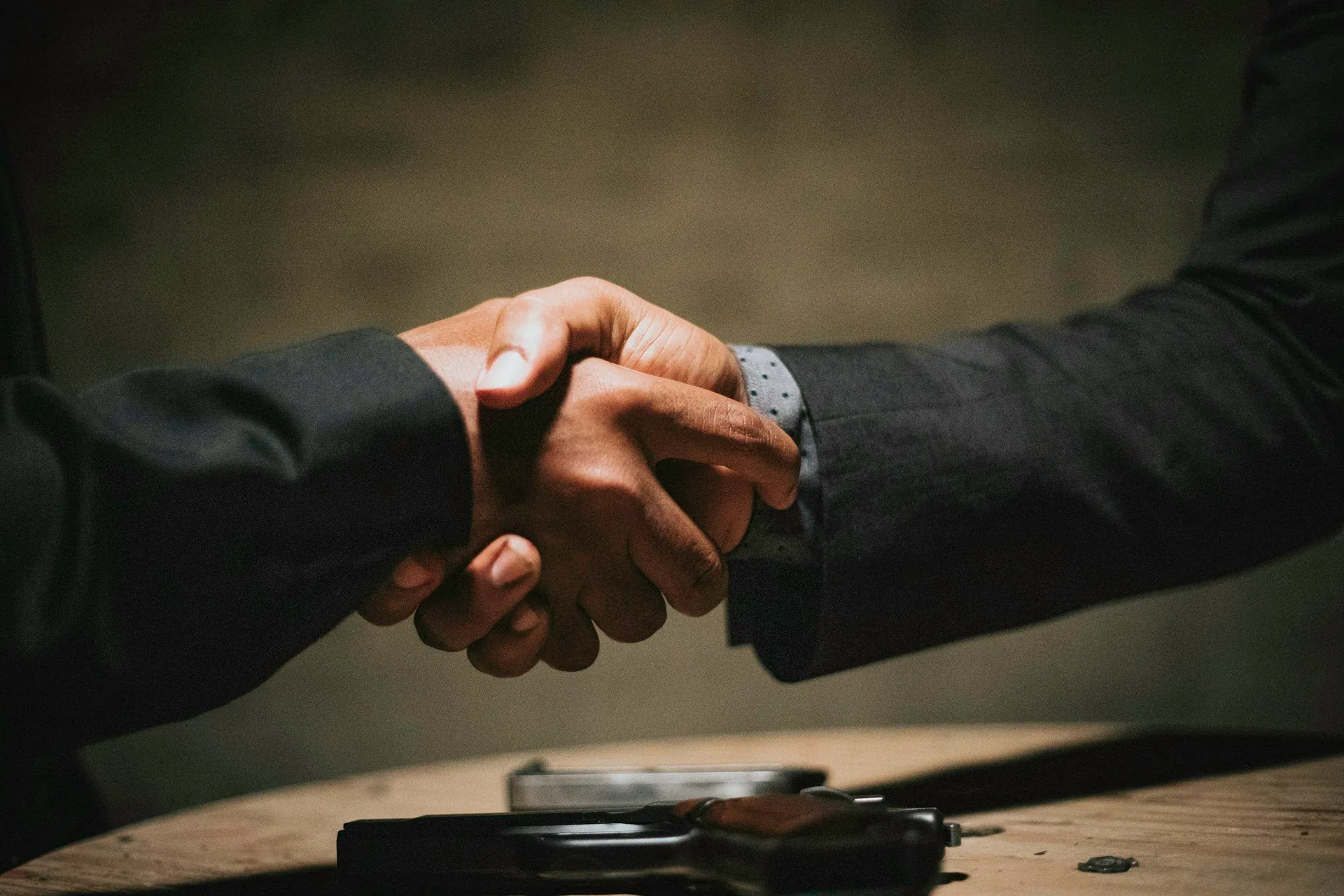 Close-up of two people shaking hands, both wearing formal suits, over a wooden table with items on it, in a professional setting.