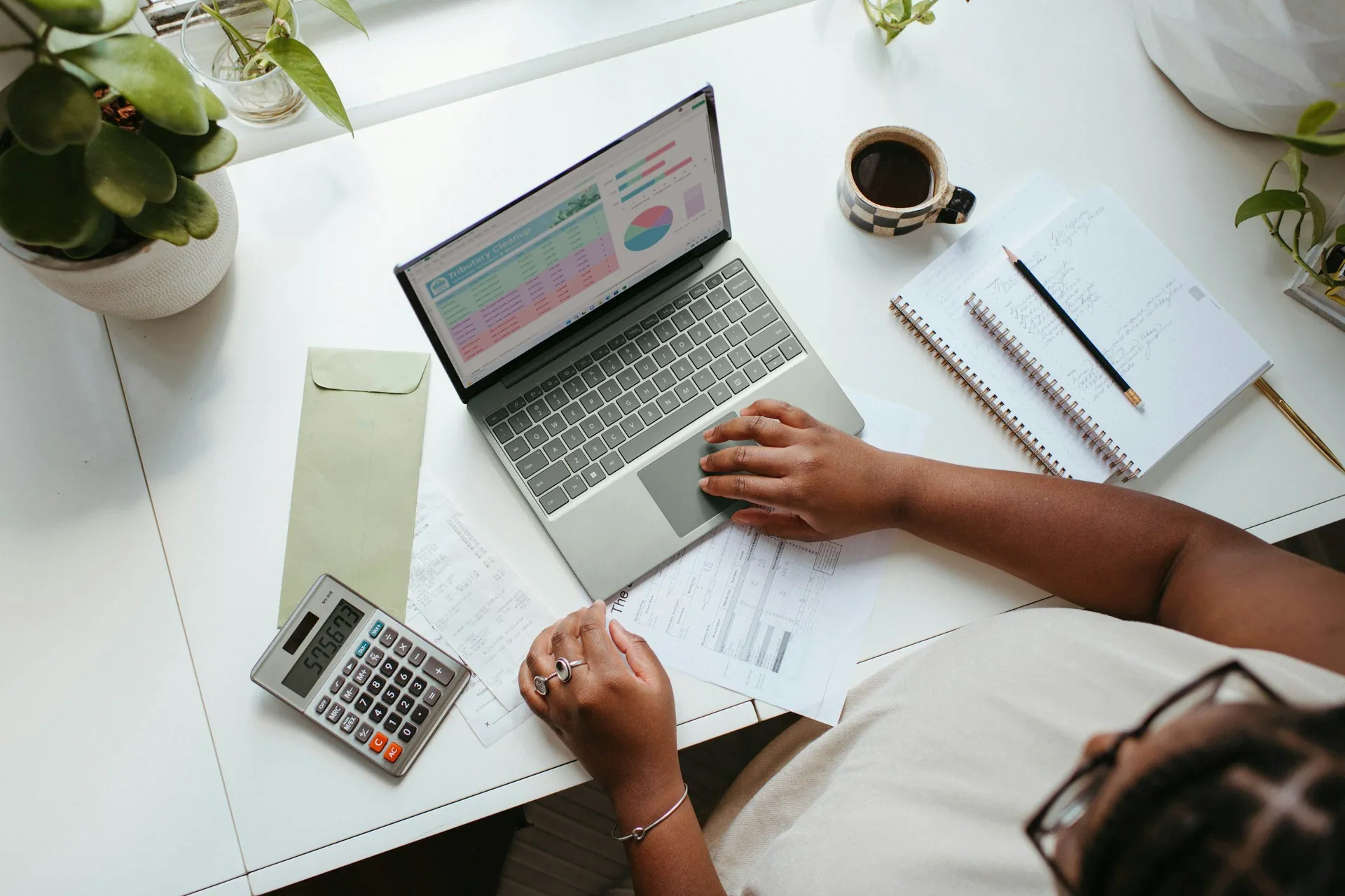 Overhead view of a person working at a white desk with a laptop, calculator, notebook, and papers, with a cup of coffee and potted plants nearby.