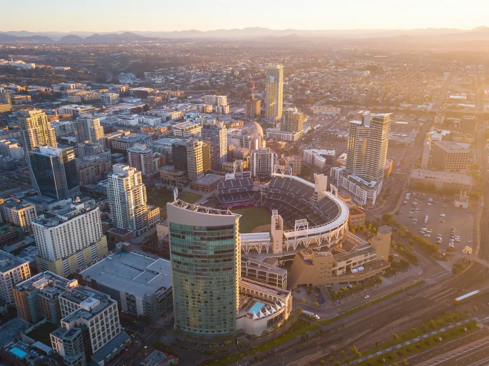 Aerial view of a city skyline at sunset showing numerous tall buildings, a stadium, and mountains in the background.