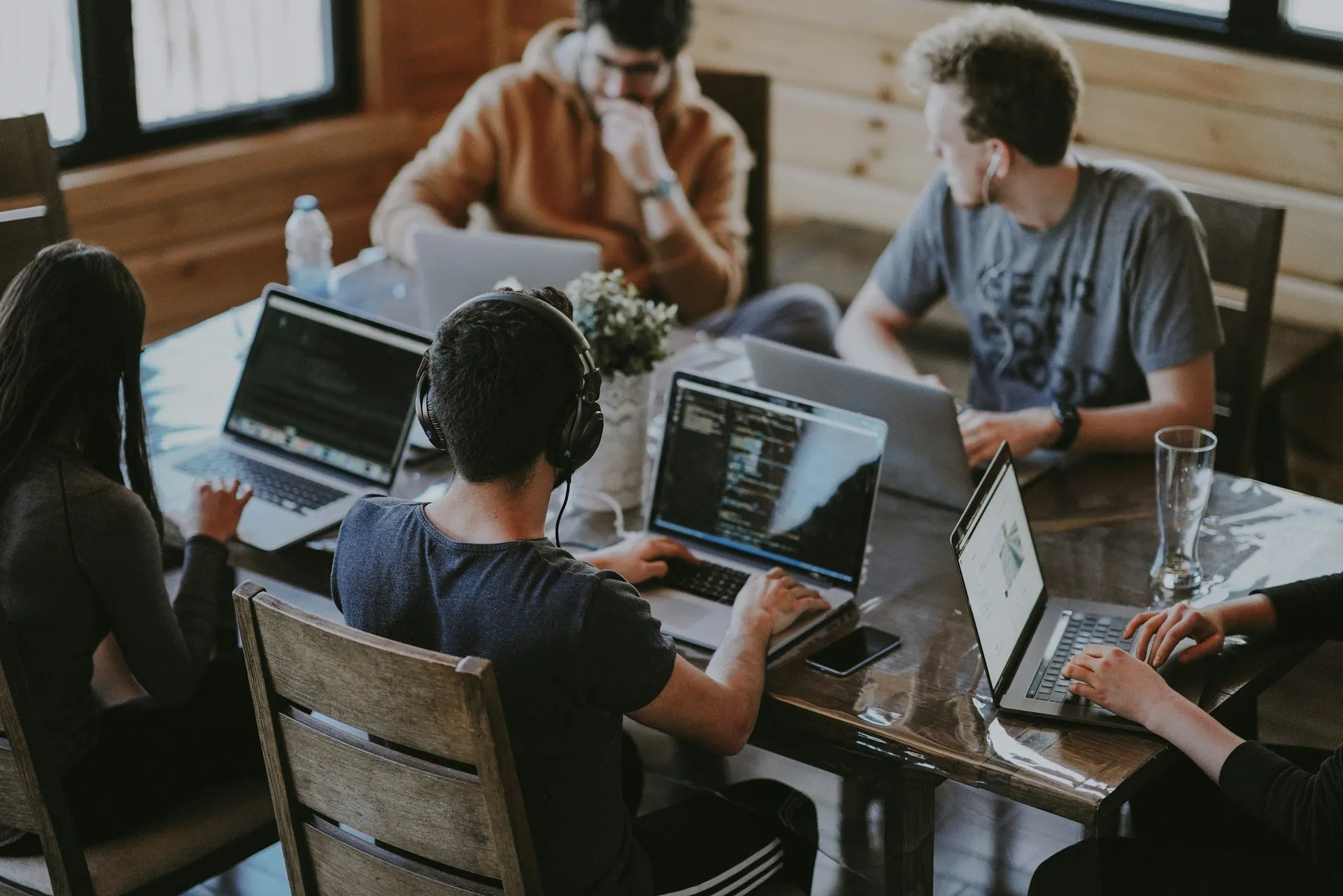Group of five young adults working on laptops and computer screens around a wooden table in a cozy, wood-paneled room.