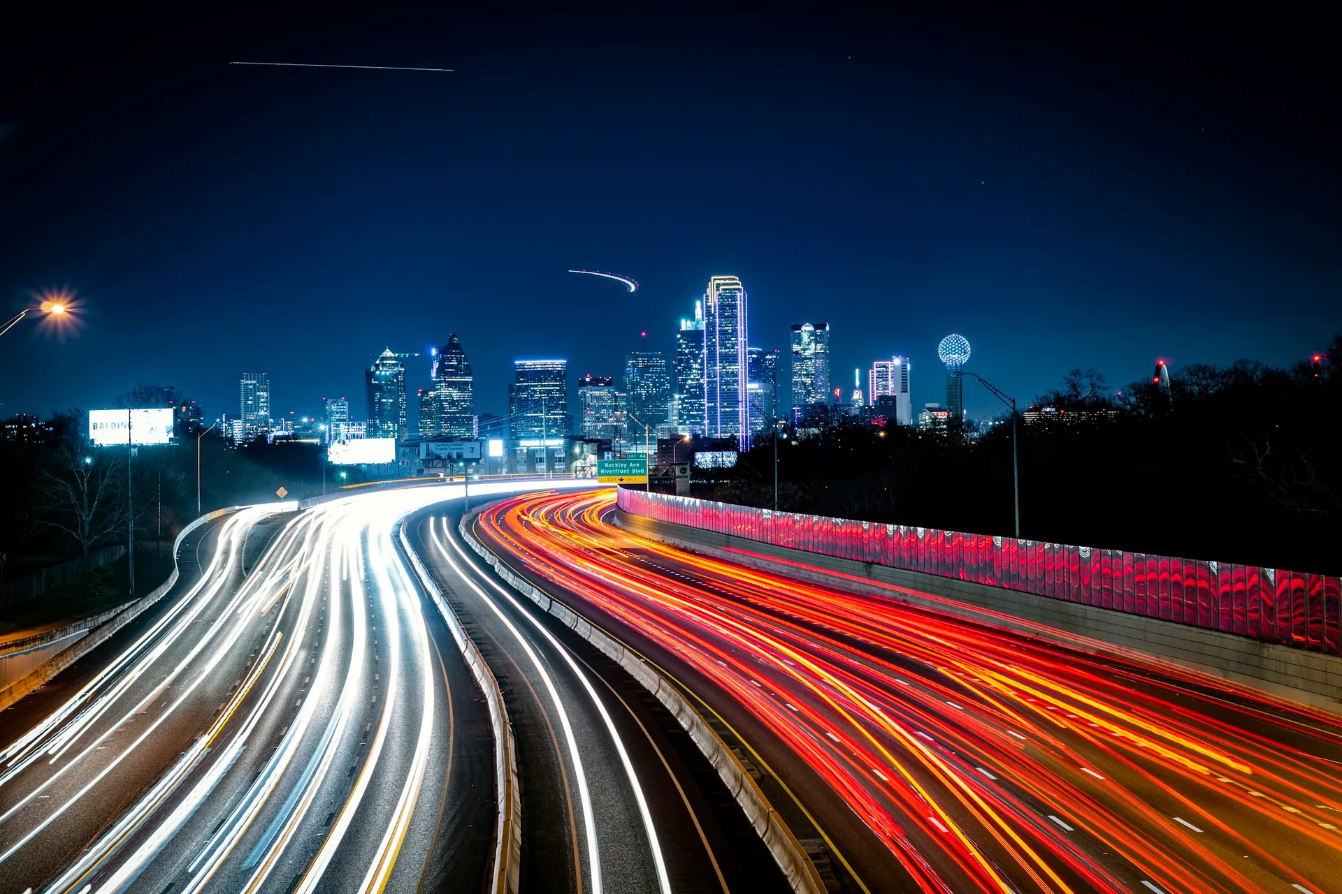 Nighttime cityscape with bright light trails from moving cars on a highway, and a city skyline with illuminated skyscrapers in the background.