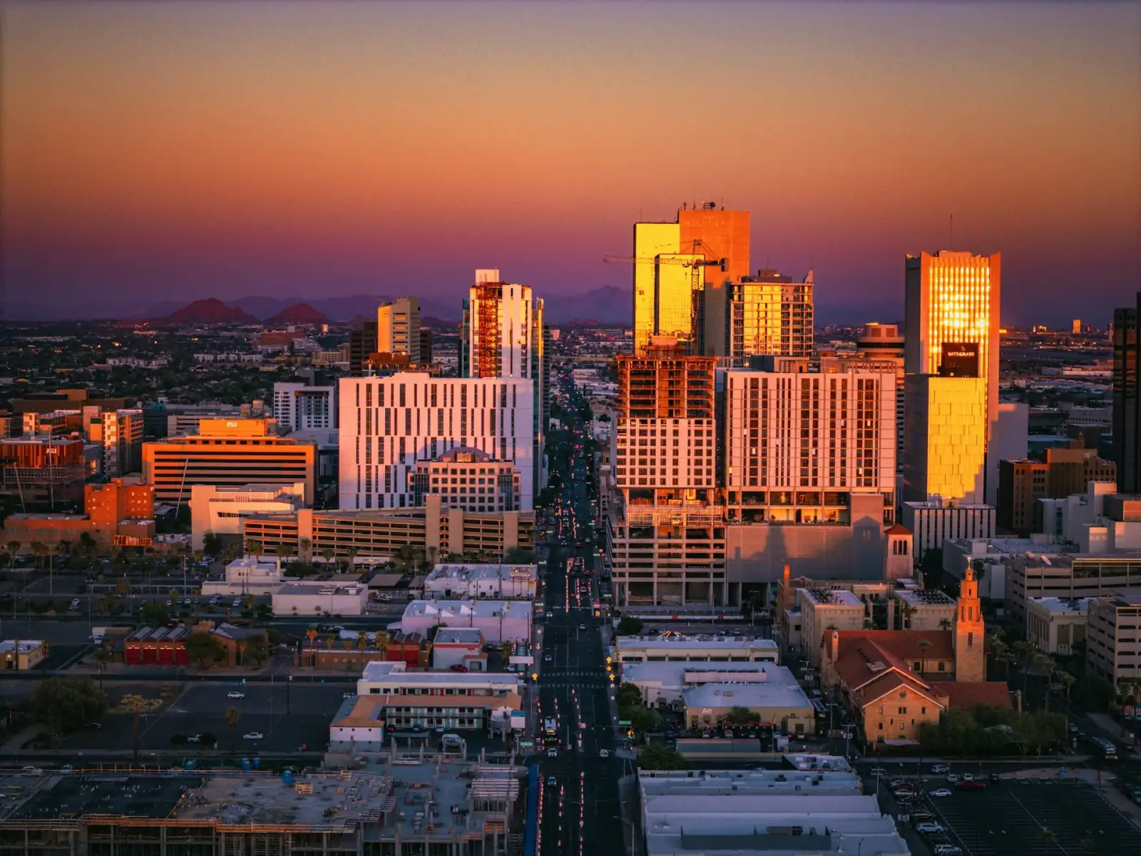 Aerial view of a city skyline during sunset with tall modern buildings reflecting orange and pink hues, and mountains in the background.