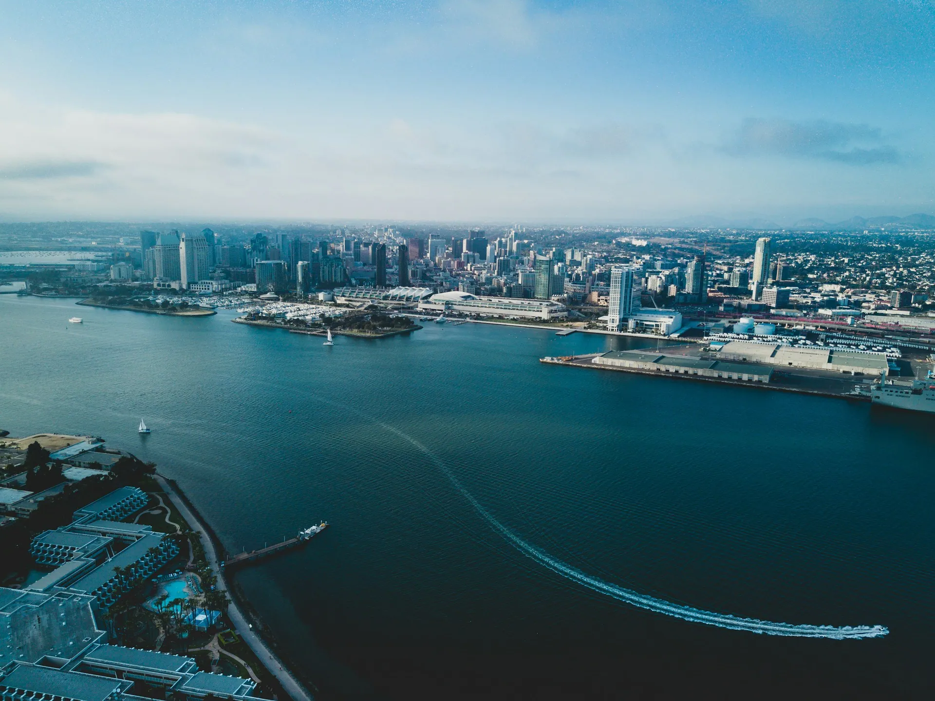 Aerial view of the city skyline, showing tall skyscrapers along the waterfront, with boats sailing on the bay and a boat leaving a wake in the water.