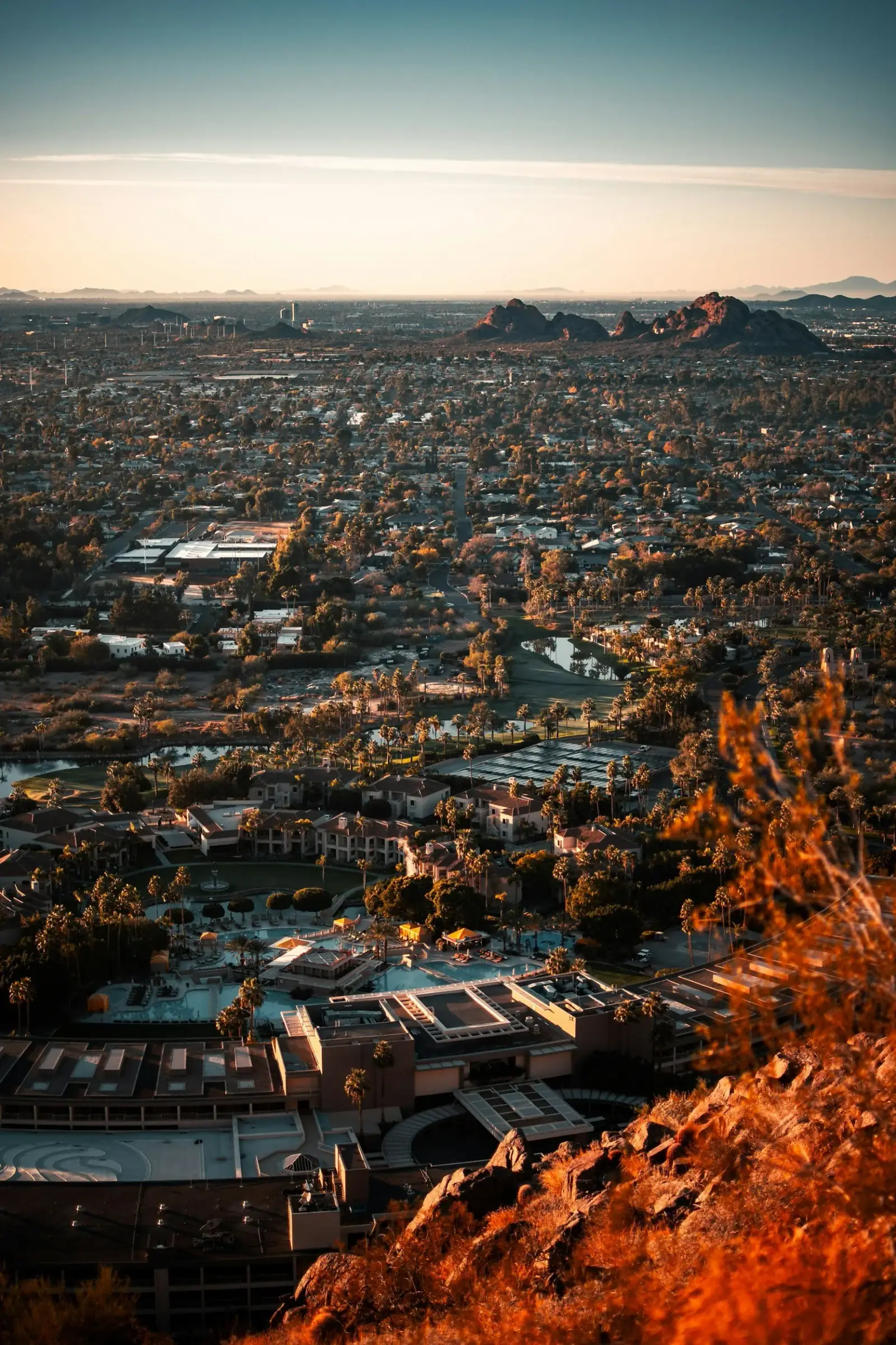 Aerial view of a cityscape at sunset, featuring residential neighborhoods, a golf course, and rocky hills in the distance.