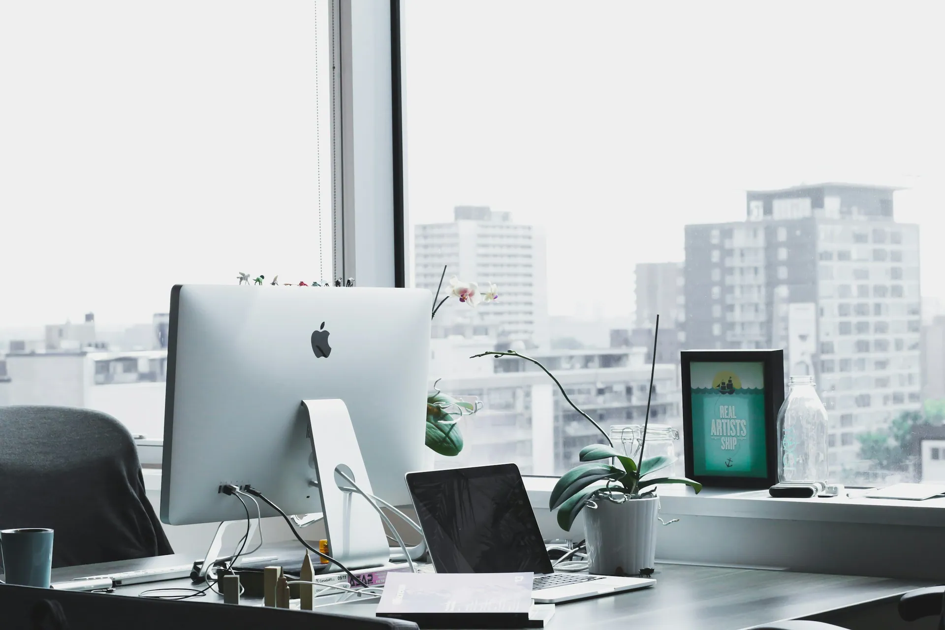 Office desk setup near a large window with a cityscape view, featuring an Apple iMac, laptop, framed artwork, potted orchid, glass water bottle, and various office supplies.