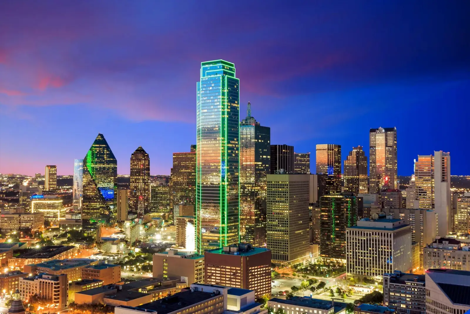 Night view of a city skyline with tall illuminated skyscrapers, including a prominent glass building with green neon outlining, under a dark blue sky with some pink clouds.